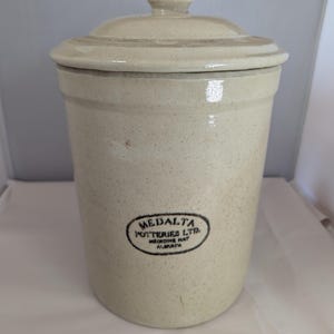 May include: A cylindrical off-white stoneware crock with a matching lid. The exterior features a speckled finish and a black oval stamp that reads Medalta Potteries Ltd. Medicine Hat Alberta. The vessel has a smooth, glazed surface and a simple knob.