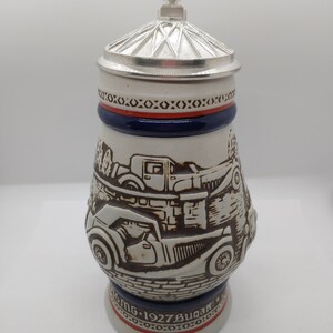 May include: A white ceramic beer stein with a silver lid. The stein features a detailed illustration of vintage cars, including a Bugatti, and the text "MG 1927 Bugatti".