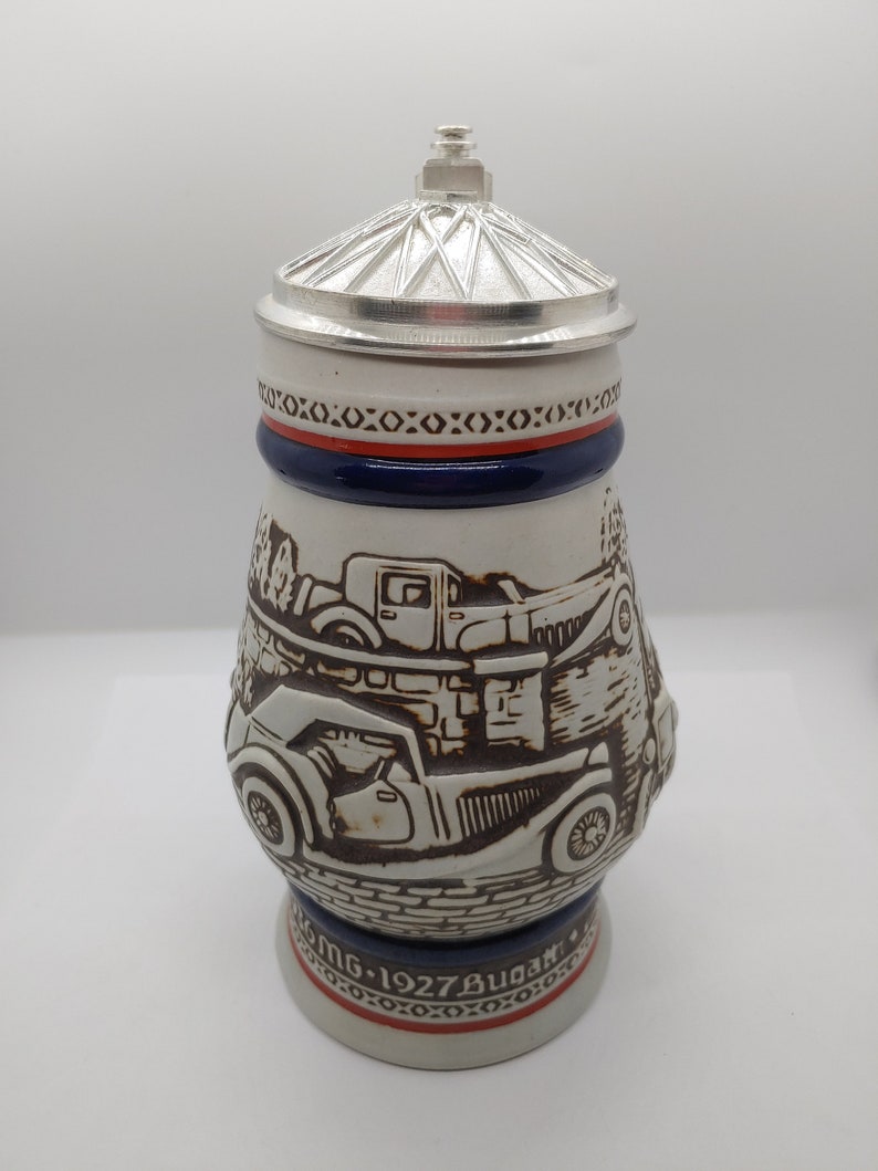 May include: A white ceramic beer stein with a silver lid. The stein features a detailed illustration of vintage cars, including a Bugatti, and the text "MG 1927 Bugatti".