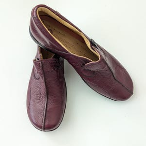 May include: A pair of burgundy leather slip-on shoes. The shoes have a textured surface and a rounded toe. The interior is a lighter shade of brown. The brand name "structured Clarks" is visible on the insole.