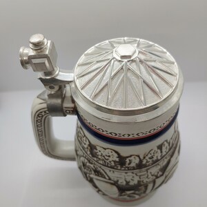May include: A white ceramic beer stein with a silver lid and a detailed design of a scene with trees and people. The lid has a starburst pattern.