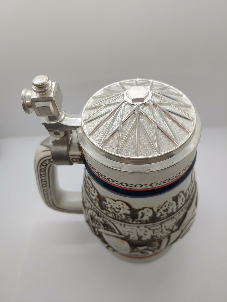 May include: A white ceramic beer stein with a silver lid and a detailed design of a scene with trees and people. The lid has a starburst pattern.