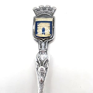 May include: A silver-colored souvenir spoon featuring a detailed design. The spoon's handle has a deer head motif, and the top displays a crest with the text "ARC TRIOMPHE" and an image of the Arc de Triomphe in blue and gold.