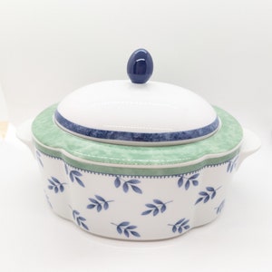 May include: A white ceramic serving dish with a green and blue floral pattern. The dish has a lid with a blue knob.