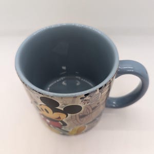 Op de afbeelding: Een keramische mok met een blauw interieur en handvat. De buitenkant heeft een vintage-stijl ontwerp met Mickey Mouse in een rood shirt en gele schoenen. De mok heeft een lichtblauwe rand en een witte achtergrond.