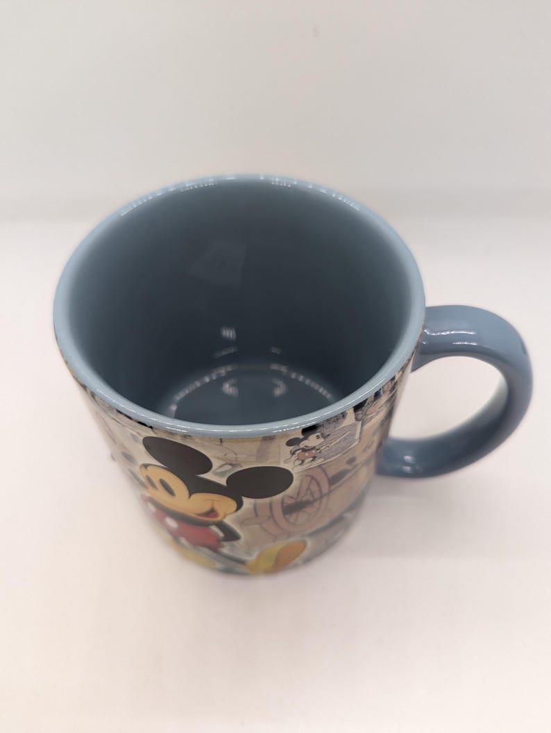 Op de afbeelding: Een keramische mok met een blauw interieur en handvat. De buitenkant heeft een vintage-stijl ontwerp met Mickey Mouse in een rood shirt en gele schoenen. De mok heeft een lichtblauwe rand en een witte achtergrond.