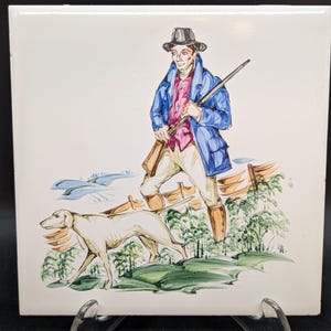 Puede incluir: Azulejo de cerámica cuadrado con una ilustración pintada a mano de un cazador con abrigo azul y camisa rosa, sosteniendo un rifle, con un perro. El fondo incluye una valla y árboles.