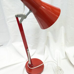 K&ouml;nnte beinhalten: Rote Schreibtischlampe mit konischem Schirm und verstellbarem Arm. Die Lampe hat einen runden Sockel und ein wei&szlig;es Netzkabel. Der Metallarm ist mit silbernen Scharnieren an Sockel und Schirm befestigt. Die Lampe ist f&uuml;r den Schreibtischgebrauch konzipiert.