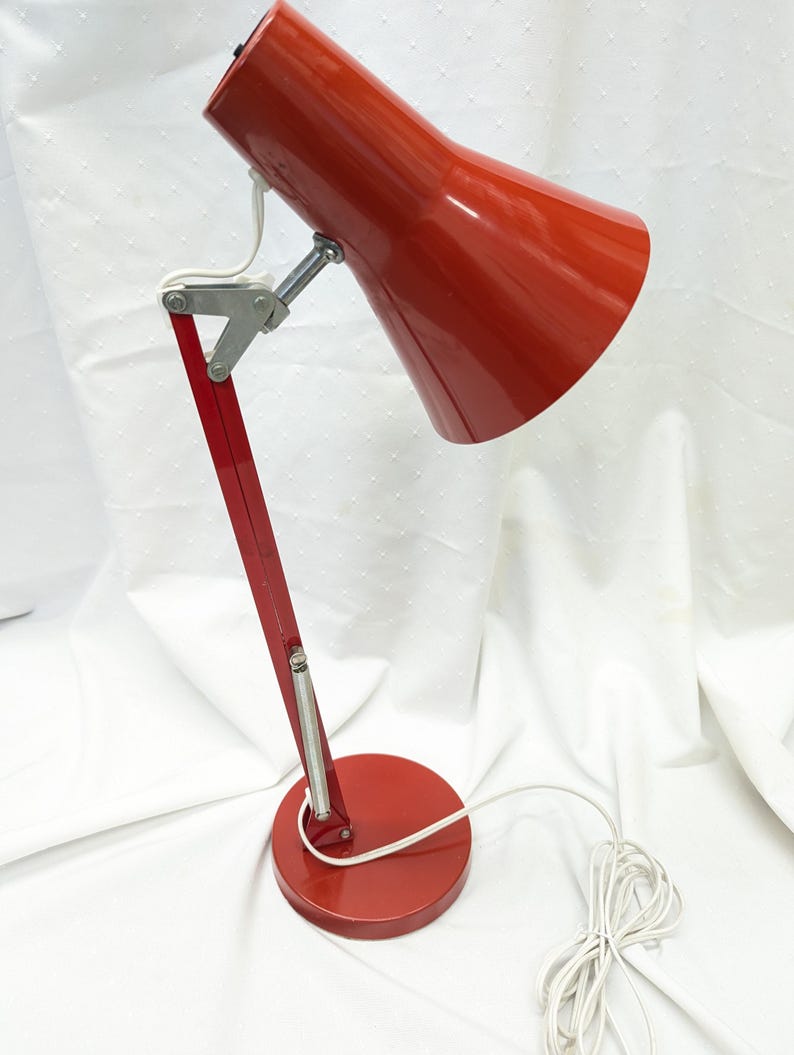 K&ouml;nnte beinhalten: Rote Schreibtischlampe mit konischem Schirm und verstellbarem Arm. Die Lampe hat einen runden Sockel und ein wei&szlig;es Netzkabel. Der Metallarm ist mit silbernen Scharnieren an Sockel und Schirm befestigt. Die Lampe ist f&uuml;r den Schreibtischgebrauch konzipiert.