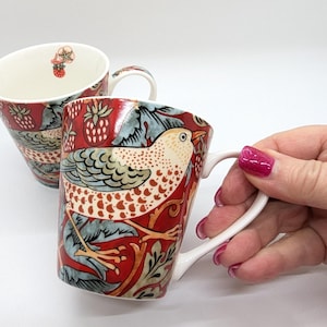 Può includere: Due tazze in ceramica rossa e bianca con un motivo di uccello e fiori. Le tazze hanno un interno bianco e un manico bianco.