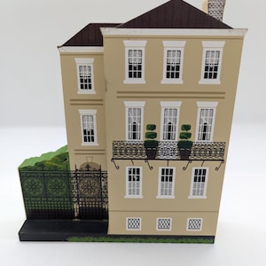 May include: A miniature beige building with a brown roof, white window frames, and a black wrought iron balcony. The building has a green fence and hedge on the side. The windows have white curtains.