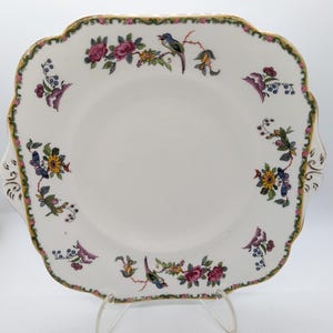 May include: Square white ceramic serving plate with a scalloped edge, featuring floral and bird designs in pink, blue, yellow, and green. The rim is trimmed with gold and green accents. The plate has two small handles.