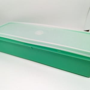 May include: A rectangular, green plastic container with a matching lid. The lid is a lighter shade of green and has a clear, circular indentation in the center. The container is designed for food storage.