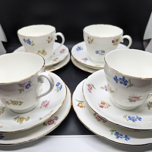 Op de afbeelding: Set van vier vintage kopjes en schotels. De witte porseleinen kopjes en schotels hebben een bloemmotief in blauw, roze en geel. Elk kopje heeft een gouden rand. De schotels hebben een geschulpte rand.