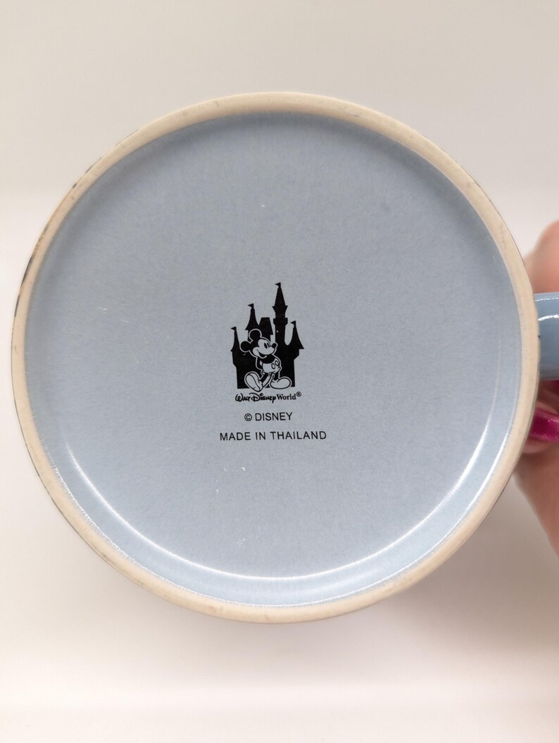 Op de afbeelding: Onderkant van een lichtblauwe keramische mok met een witte rand. De mok heeft een zwart silhouet van een kasteel met Mickey Mouse en de tekst "Walt Disney World", "&copy; Disney" en "MADE IN THAILAND".