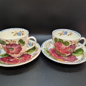 May include: Two cream-colored teacups and saucers with floral designs. The cups and saucers feature a hand-painted design of large, red and purple flowers with green leaves and blue accents. The rims are trimmed with blue.