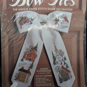 May include: A cross-stitch kit for a door decoration shaped like a bow tie. The design is titled "Winter Wonderland" and features embroidered winter scenes of houses and a church. The finished size is 11" x 17" (28 cm x 43 cm).