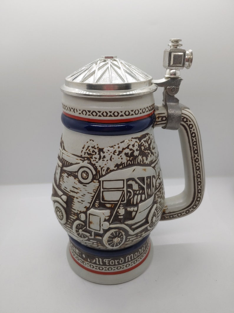 May include: A white ceramic beer stein with a brown and white image of a vintage Ford Model T car. The stein has a blue and red band around the top and a silver lid with a silver handle. The stein reads "All Ford Models".