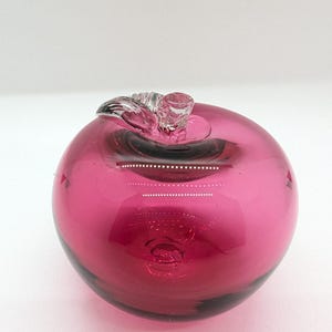 May include: A deep pink glass apple with a clear glass stem and leaf. The apple is a decorative object.