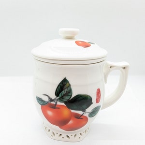 May include: White ceramic tea mug with lid, decorated with red apples and green leaves. The mug has a handle and a decorative pierced base. A small red apple is on the lid. Chinese characters are on the side.