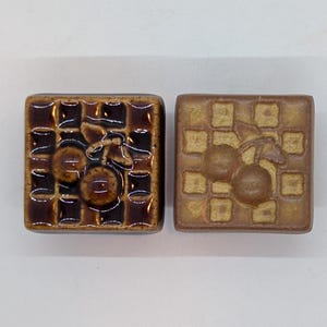 May include: Two ceramic tiles with a brown and white checkered pattern. Each tile features a raised design of a cherry with a stem and leaf.