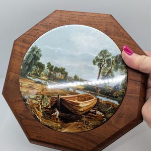 Puede incluir: Caja decorativa octogonal de madera con un inserto redondo de cerámica pintada. El inserto representa un paisaje con un barco, árboles y un río. La caja está sujeta por una mano con esmalte de uñas rosa.