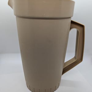 Può includere: Una brocca di plastica beige con coperchio e manico. La brocca ha un beccuccio per versare e una base scanalata. Il manico è rettangolare e attaccato al lato. Il coperchio ha un piccolo pomello.