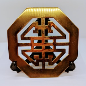 Könnte beinhalten: Eine goldfarbene, achteckige Dekorplatte aus Metall mit einem ausgeschnittenen Design eines traditionellen chinesischen Symbols. Die Platte steht auf einem schwarzen Ständer.