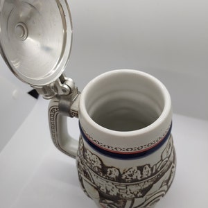 May include: A white ceramic beer stein with a brown and white scene of people and a horse-drawn carriage. The stein has a blue and red trim around the top and a silver lid.