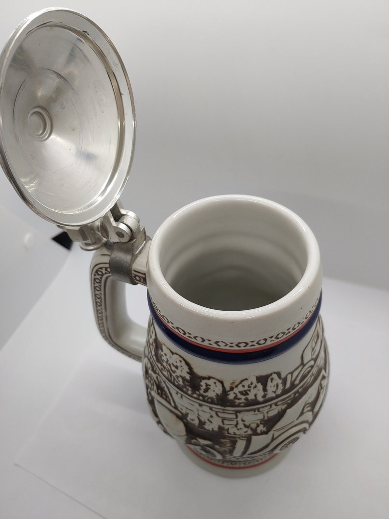 May include: A white ceramic beer stein with a brown and white scene of people and a horse-drawn carriage. The stein has a blue and red trim around the top and a silver lid.