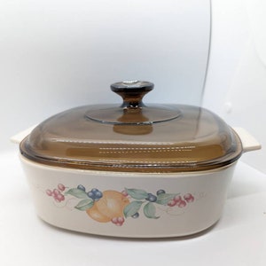 Può includere: Una casseruola in ceramica bianca con un coperchio in vetro marrone. La casseruola ha un design floreale con frutta e bacche.