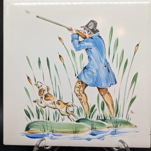 Könnte beinhalten: Quadratische Keramikfliese mit handgemalter Jagdszene. Ein Jäger in einem blauen Mantel zielt mit einem Gewehr, mit einem Hund, der vor ihm springt. Die Szene umfasst Schilf und Wasser, vor weißem Hintergrund.