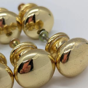 Könnte beinhalten: Vier goldfarbene Schrankknöpfe mit runder, leicht gewölbter Form. Die Knöpfe haben eine glänzende, polierte Oberfläche.