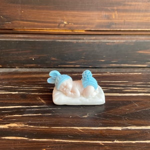 May include: A small, porcelain figurine of a sleeping baby. The baby is wearing a blue hat and diaper cover with a pom-pom. The baby is lying on a white pillow. The figurine is on a dark brown wooden surface.