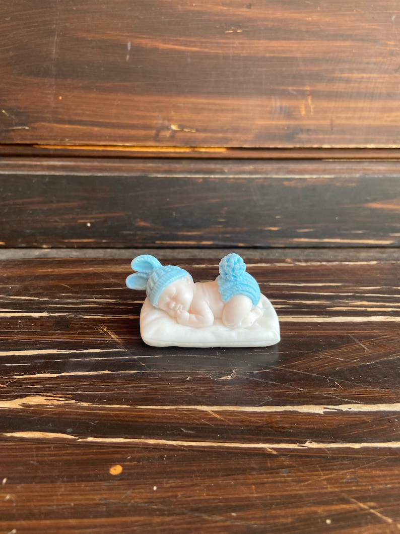 May include: A small, porcelain figurine of a sleeping baby. The baby is wearing a blue hat and diaper cover with a pom-pom. The baby is lying on a white pillow. The figurine is on a dark brown wooden surface.