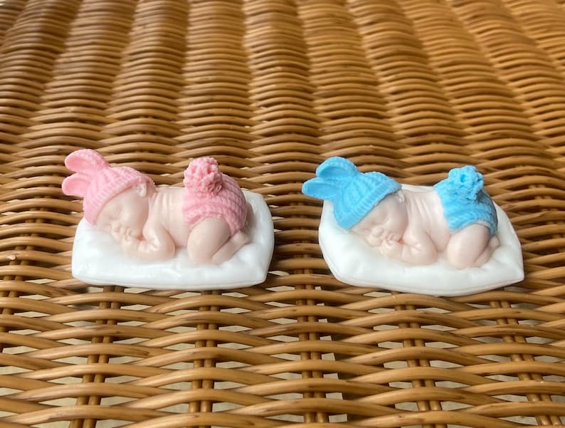 May include: Two baby figurines, one in pink and one in blue, each lying on a white pillow. The pink figurine wears a pink hat and diaper cover. The blue figurine wears a blue hat and diaper cover. Both are on a woven background.