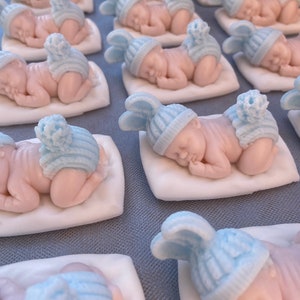 May include: Rows of baby figurines, each lying on a white pillow. The babies are light pink with blue knitted hats and diaper covers. The hats have a pom-pom on top. The figurines are arranged on a gray surface.