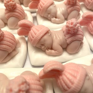 May include: A collection of sleeping baby figurines, each lying on a white pillow. The babies are light pink and wear matching pink knit hats with pom-poms or bunny ears. The figurines are likely for a baby shower or nursery decor.