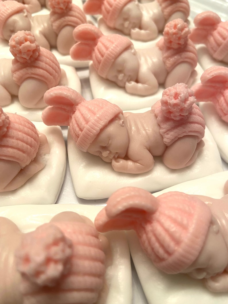 May include: A collection of sleeping baby figurines, each lying on a white pillow. The babies are light pink and wear matching pink knit hats with pom-poms or bunny ears. The figurines are likely for a baby shower or nursery decor.