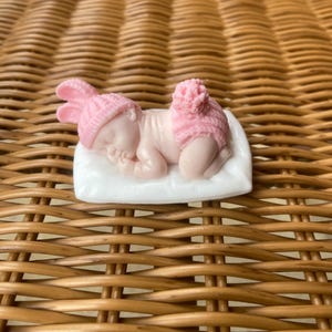 May include: A small, pink figurine of a sleeping baby wearing a bunny hat and diaper cover. The baby is lying on a white pillow. The figurine is on a woven, brown surface.