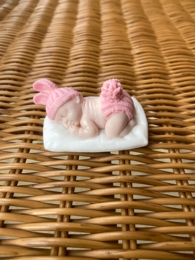 May include: A small, pink figurine of a sleeping baby wearing a bunny hat and diaper cover. The baby is lying on a white pillow. The figurine is on a woven, brown surface.