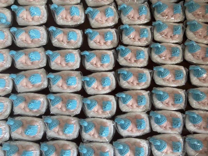 May include: Rows of baby-shaped candies, each wrapped in clear plastic. The candies are pink with blue hats. The candies are arranged in a grid pattern, creating a visually appealing display.