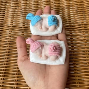 May include: Two baby-shaped soaps, one with a blue hat and diaper, the other with pink. Each soap rests on a white rectangular base. The soaps are held in a hand, with a woven background.