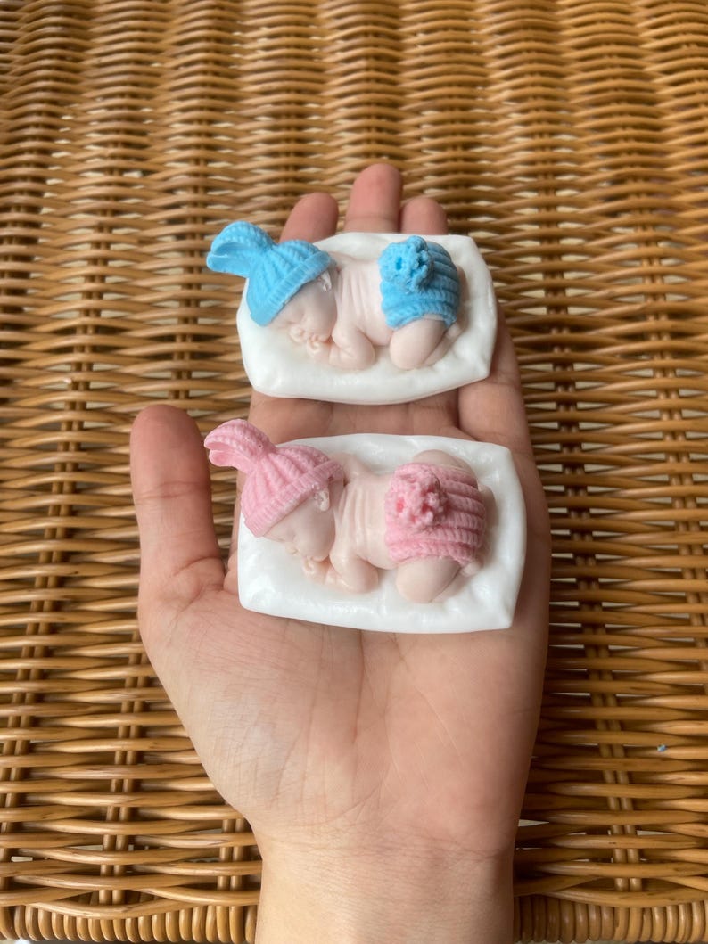 May include: Two baby-shaped soaps, one with a blue hat and diaper, the other with pink. Each soap rests on a white rectangular base. The soaps are held in a hand, with a woven background.