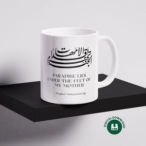 Könnte beinhalten: Weiße Keramik-Tasse mit schwarzer arabischer Kalligraphie und dem englischen Text „PARADISE LIES UNDER THE FEET OF MY MOTHER - Prophet Muhammad“. Die Tasse steht auf einem schwarzen Regal, mit einem digitalen Download-Symbol.