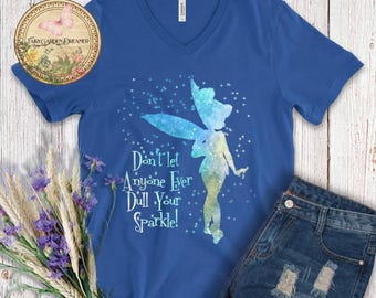 May include: A blue V-neck t-shirt featuring a fairy silhouette with blue and yellow watercolor wings and a starry background. The text "Don't let Anyone Ever Dull Your Sparkle!" is printed below the fairy. Denim shorts are visible.