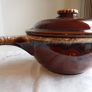 May include: A brown glazed ceramic casserole dish with a lid and a handle. The dish has a white glaze on the handle and rim.