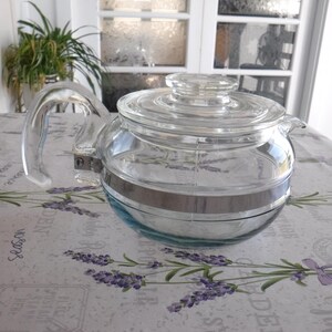 May include: A clear glass teapot with a matching glass lid and a curved glass handle. The body of the teapot features a horizontal silver-toned metal band that encircles the middle section, connecting to the handle with a metal bracket.