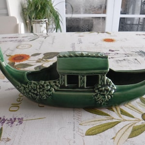 May include: A green ceramic gondola-shaped planter. The planter features a detailed design with a cabin and floral accents. The gondola is approximately 46 cm long, perfect for displaying small plants or decorative items.