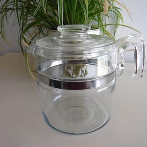 May include: A clear glass coffee percolator pot featuring a matching glass lid and a sturdy glass handle. The cylindrical body is accented with a polished silver-tone metal band encircling the centre, providing a clean and functional design.